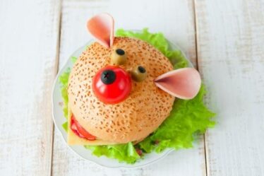 A sandwich on a plate is decorated to look like a bear’s face, complete with olives for eyes and a cherry tomato nose. With fresh green lettuce and protein-rich cooked meat, it’s a fun and healthy meal option sitting on a white wooden surface.