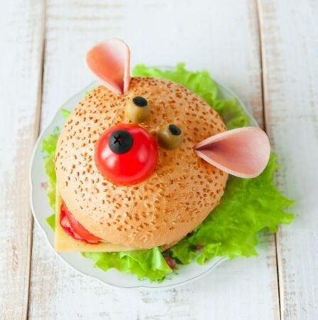 A sandwich on a plate is decorated to look like a bear’s face, complete with olives for eyes and a cherry tomato nose. With fresh green lettuce and protein-rich cooked meat, it’s a fun and healthy meal option sitting on a white wooden surface.