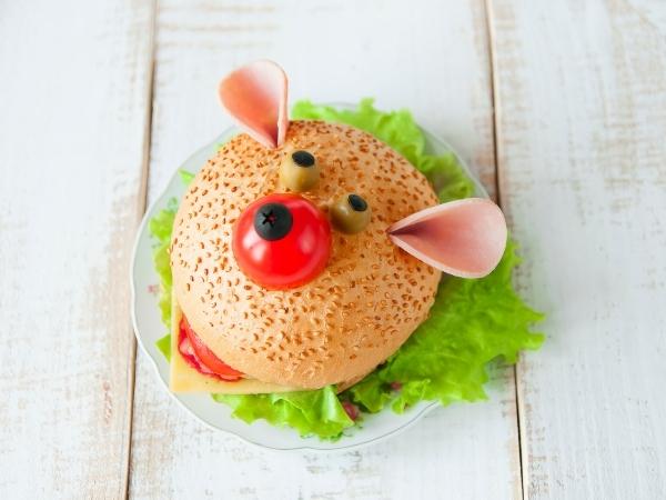 A sandwich on a plate is decorated to look like a bear’s face, complete with olives for eyes and a cherry tomato nose. With fresh green lettuce and protein-rich cooked meat, it’s a fun and healthy meal option sitting on a white wooden surface.