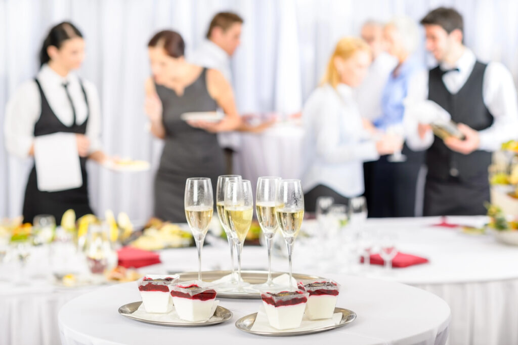 A close-up of a table with four champagne flutes and desserts in glass cups with red topping. In the blurred background at a wedding venue, people in formal attire and servers converse amid white tablecloths and elegant décor.