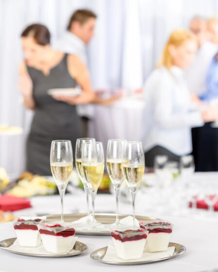 A close-up of a table with four champagne flutes and desserts in glass cups with red topping. In the blurred background at a wedding venue, people in formal attire and servers converse amid white tablecloths and elegant décor.