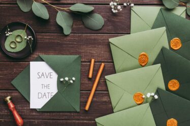 A “Save the Date” card peeks from a green envelope on a wooden table. Surrounding it are more green envelopes sealed with wax—perfect for couples seeking simple wedding hacks to save money on beautiful, budget-friendly invitations.