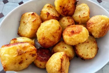 A white oval plate filled with golden-brown, crispy roast potatoes sits on a patterned surface. The potatoes have slightly rough, crunchy exteriors with some darker, caramelised spots and appear fluffy and soft inside—just like the perfect results from a classic roast potatoes recipe.