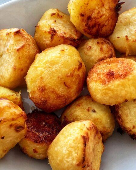 A white oval plate filled with golden-brown, crispy roast potatoes sits on a patterned surface. The potatoes have slightly rough, crunchy exteriors with some darker, caramelised spots and appear fluffy and soft inside—just like the perfect results from a classic roast potatoes recipe.